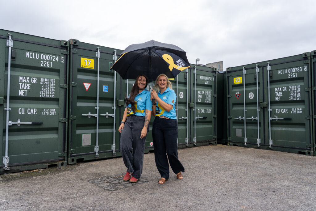 Two team members sheltering under an umbrella between rows of storage containers