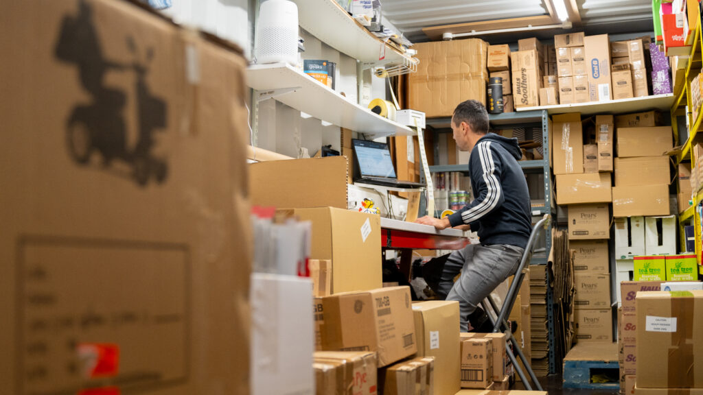Online retailer packing orders at a desk surrounded by stock in a business pod